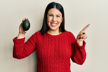 Sticker - Beautiful brunette woman holding avocado smiling happy pointing with hand and finger to the side