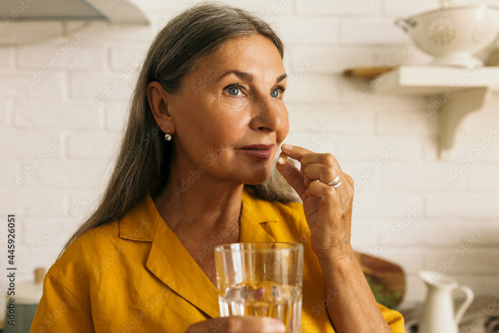 Cute mature female holding drugs and glass of water in hands. Elderly woman taking medication. Treatment course or vitamins complex idea. Healthy lifestyle and healthcare concept - obrazy, fototapety, plakaty 