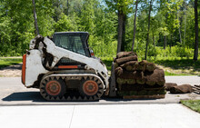 A Compact Front Loader And A Pallet With Rolls Of Sod Grass For Installation At A New Home Construction Work Site, With Trees In The Background And Concrete In The Foreground.