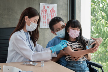 Asian girl taking selfie portrait with mobile phone with father while a doctor is vaccinating prevent coronavirus (COVID-19) at hospital clinic. Injections treat fever and illness with medication.