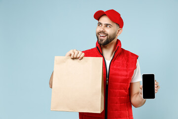 Wall Mural - Delivery guy employee man in red cap white T-shirt vest uniform work as dealer courier service hold brown clear blank craft paper takeaway bag use cell phone isolated on pastel blue background studio.