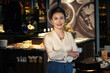 © DragonImages - Portrait of happy smiling elegant middle-aged woman standing at bar counter of restaurant she is opening