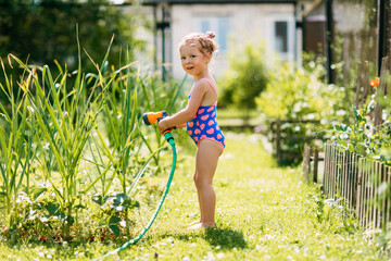 Charming baby girl waters plants with a hose in the garden in the backyard of the house on a sunny summer day