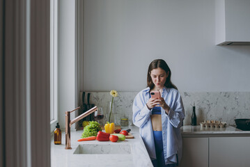 Wall Mural - Young pensive housewife woman 20s wearing casual clothes blue shirt prepare salad use mobile cell phone look for online recipe cook food in light kitchen at home alone Healthy diet lifestyle concept.