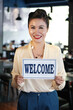 © DragonImages - Portrait of happy smiling restaurant owner holding welcome sign in her hands and looking at camera