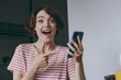 Young surprised housewife woman in casual clothes striped t-shirt point index finger on mobile cell phone chatting online browsing internet in light kitchen at home alone People lifestyle concept.