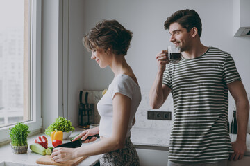 Young happy couple two caucasian woman man 20s in casual t-shirt clothes drink coffee in morning prepare vegetable salad cooking food in light kitchen at home together. Healthy diet lifestyle concept.