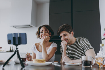 Wall Mural - Young confused fun couple two woman man in casual clothes sit by table eat pancake watch live stream by mobile cell phone prop up chin cook food in light kitchen at home together Healthy diet concept.