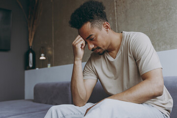 Wall Mural - Side view pensive thoughtful confused young african american man in beige t-shirt sit on grey sofa indoors apartment procrastinate prop up forehead rest on weekends stay at home Tattoo translate fun.