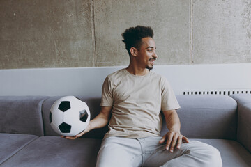 Wall Mural - Young african american man football fan in beige t-shirt cheer up support favorite team with soccer ball sit on grey sofa indoors apartment look aside People sport leisure stay home lifestyle concept.