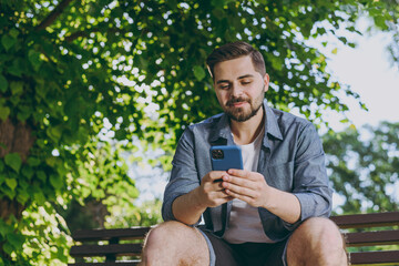 Wall Mural - Bottom view smiling happy fun nyoung man in blue shirt sit on bench use mobile cell phone chat online rest relax in spring green city sunshine park outdoors on nature Urban lifestyle leisure concept