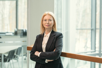 Wall Mural - Portrait of confident mature businesswoman in suit standing with her arms crossed in the office corridor and looking at camera