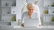© AboutLife - Senior lady secretary with grey hair and glasses in elegant suit types on laptop at table against designed bookshelves