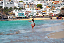 View Of The White Buildings On The Rocky Shore Of Morro Jable And A Child Running On The Beach With Turquoise Blue Ocean Water. Fuerteventra, Canary Islands, Spain