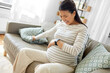 © Syda Productions - pregnancy, rest, people and expectation concept - pregnant asian woman with smart watch sitting on sofa at home having labor contractions pain