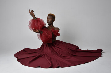 Wall Mural - Full length portrait of pretty African woman wearing long red renaissance medieval fantasy gown, sitting pose on a light grey studio background.