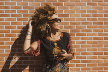 Beautiful African American Woman Leaning Against Orange Bricks Wall Wearing Earphones Listening To Music On Smartphone And Smiling. Copy Space For Text.