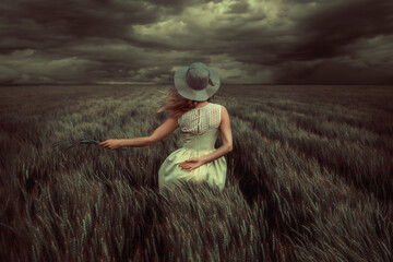 Young blonde woman standing in a wheat field, wearing a yellow summer dress, on a windy and stormy day