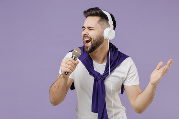 Wall Mural - Happy charismatic young brunet man 20s wears white t-shirt purple shirt listen to music in headphones keep eyes closed hand up sing in microphone isolated on pastel violet background studio portrait