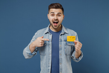 Shocked surprised happy excited young brunet man 20s wear denim jacket hold in hand credit bank card pointing on it isolated on dark blue background studio portrait. Achievement career wealth concept
