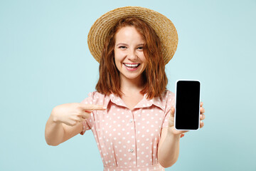 Young redhead woman 20s hold mobile cell phone with blank screen workspace area point on it with finger wear casual dress straw hat look camera isolated on pastel blue color background studio portrait