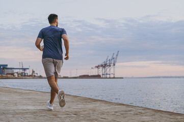 Full body back view young strong sporty athletic toned fit sportsman man in sports clothes warm up training run jog at sunrise sun over sea beach outdoor on pier seaside in summer day cloudy morning