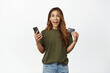 Online shopping. Excited smiling middle aged woman, holding smartphone, showing credit discount card, talking about big sale, standing against white background