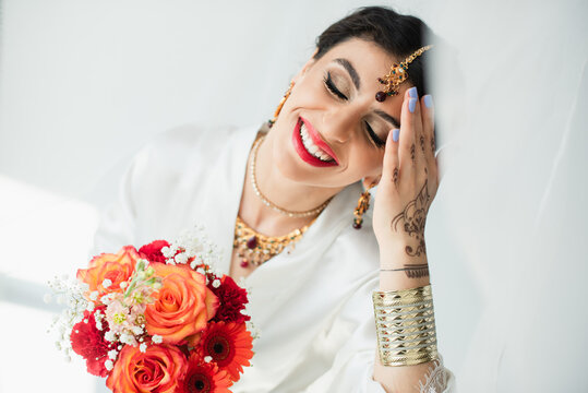 young joyful indian bride with mehndi holding bouquet of flowers on white