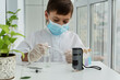 © Anna - Boy in white medical robe, mask and gloves holds pipette and glass and preparing to look into little microscope. Scientifil experiments of young scientist.
