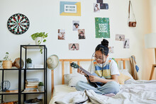 Full Length Portrait Of Asian Teenage Girl Playing Ukulele While Sitting On Bed In Cozy Room, Copy Space