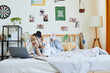 © Seventyfour - Portrait of teenage Asian girl using laptop on bed in cozy room interior, copy space