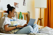 © Seventyfour - Side view portrait of teenage Asian girl using laptop while sitting on bed in cozy room interior, copy space