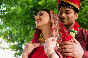 Wall Mural - low angle view of pleased indian man in turban hugging bride in red sari