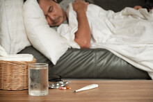Asian man is absent from work and sick leave because of illness, takes temperature by thermometer, takes medicine, and sleeps in blankets on sofa at home, tissues, and capsule pills on the table.