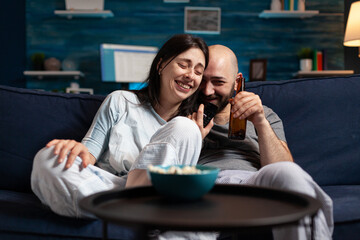 Wall Mural - Relaxed couple in pajamas relaxing on sofa eating popcorn watching comedy movie, enjoying free time together. Husband and wife relaxing late at night in living room. Looking at entertainment film