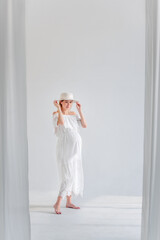 Wall Mural - Young pregnant woman in anticipation of a baby in a white hat, dress, hugs her belly. Tender blonde girl in a minimalistic, isolated interior of airy fabrics. Happy maternity pregnancy, surrogacy