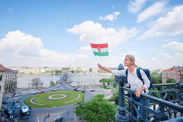 Wall Mural - Enjoying vacation in Budapest. Young traveling woman with national hungarian flag walking on riverside promenade with city view.