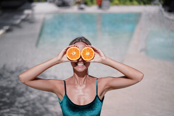 Wall Mural - Outdoor portrait of a lovely young woman standing over swimming pool showing slices of oranges.