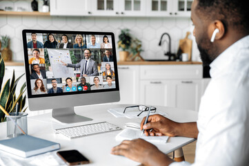Wall Mural - Online education, business seminar. African American smart confident man sit at home at work desk, uses computer, listening to an online financial lecture, taking notes, diverse people on the screen