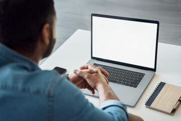 Rear view of young man freelance sitting at comfortable workplace and using laptop computer. Business man watching online training webinar, having videoconference meeting on white mockup screen