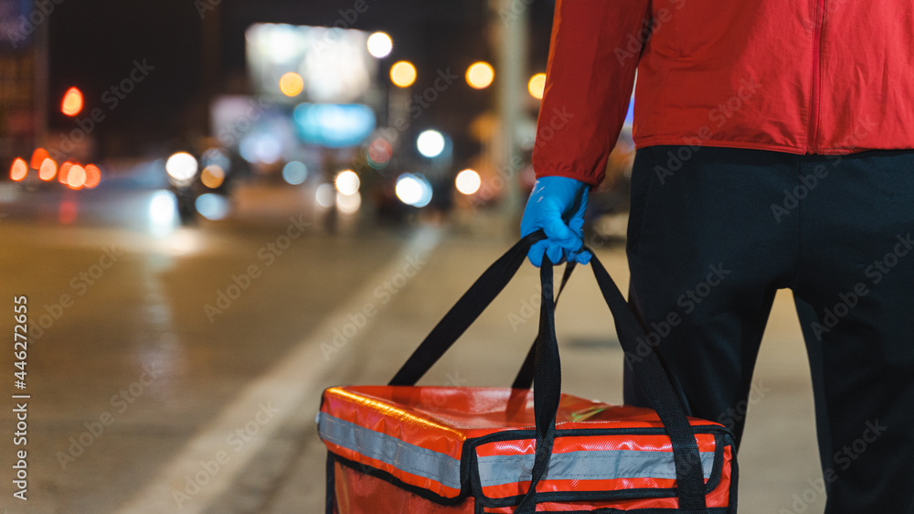 Close-up shot of a food deliveryman in red uniform carrying a food delivery box to deliver for customer for order during COVID-19 outbreak lockdown in the city at night time - obrazy, fototapety, plakaty 
