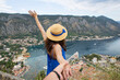 A girl in a blue dress and a straw hat held by a musky hand stands against the background of Boka Kotorska Bay on a beautiful summer day, Montenegro.