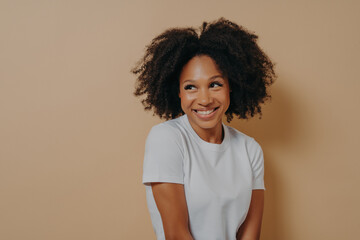 Wall Mural - Cute african woman with toothy smile looking aside with shyness, isolated over beige background