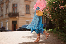 Summer Street Fashion Details: Elegant Woman Wearing Trendy Pink Shirt With Knot, Polka Dot Blue Midi Skirt, White Strap Sandals, With Yellow Wicker Leather Shoulder Bag, Walking In Street. Copy Space