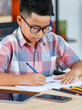 © Bangkok Click Studio - Smart Asian boy wearing glasses and grey checked shirt sitting on studying desk of tablet, mobile phone, and colorful pencils in reading room and concentrate on drawing on white paper