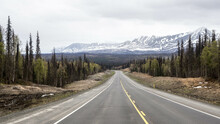 Empty Long Highway Through Pine Forest With Snowy Mountains In The Background In Winter