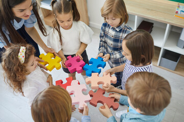 Team of little school children with teacher standing in circle and joining colorful jigsaw puzzle pieces trying to fit them together, high angle close up. Education, fun, teamwork in classroom concept
