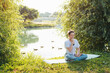Young woman meditating by the lake, performing alternating nostril breathing, outdoors. Woman doing yoga asana nadi shodhana pranayama at sunrise in the park. Yoga and meditation in nature.