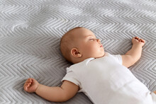Cute Newborn Baby Wearing White Bodysuit Lying On Bed On Grey Blanket, Charming Baby Relaxing At Home After Walking.