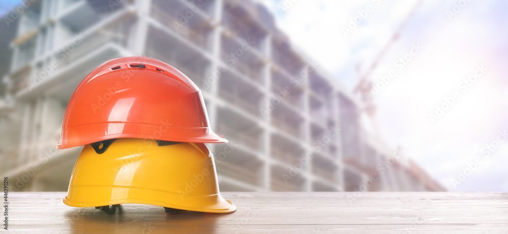 Hard hats on wooden surface at construction site with unfinished building, banner design. Space for text - obrazy, fototapety, plakaty 